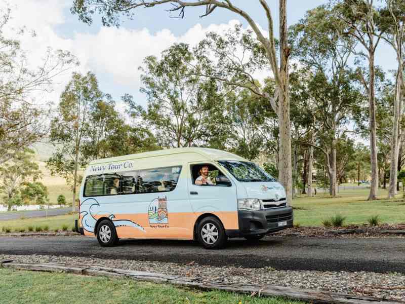 Vintage-style tour van parked on a scenic road surrounded by lush trees, promoting Newy Tour Co. for Newcastle and Hunter Valley excursions.