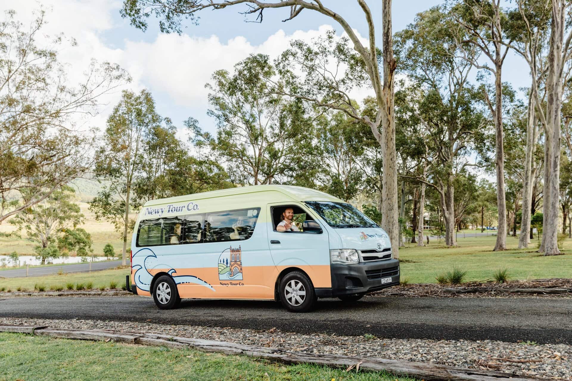 Vintage-style tour van parked on a scenic road surrounded by lush trees, promoting Newy Tour Co. for Newcastle and Hunter Valley excursions.