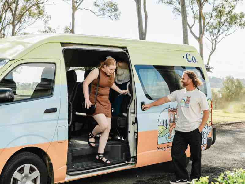 Comfortable Newy Tour Co. van with scenic outdoor backdrop, featuring friendly guide helping travelers, offering sightseeing tours in Newcastle, Australia.
