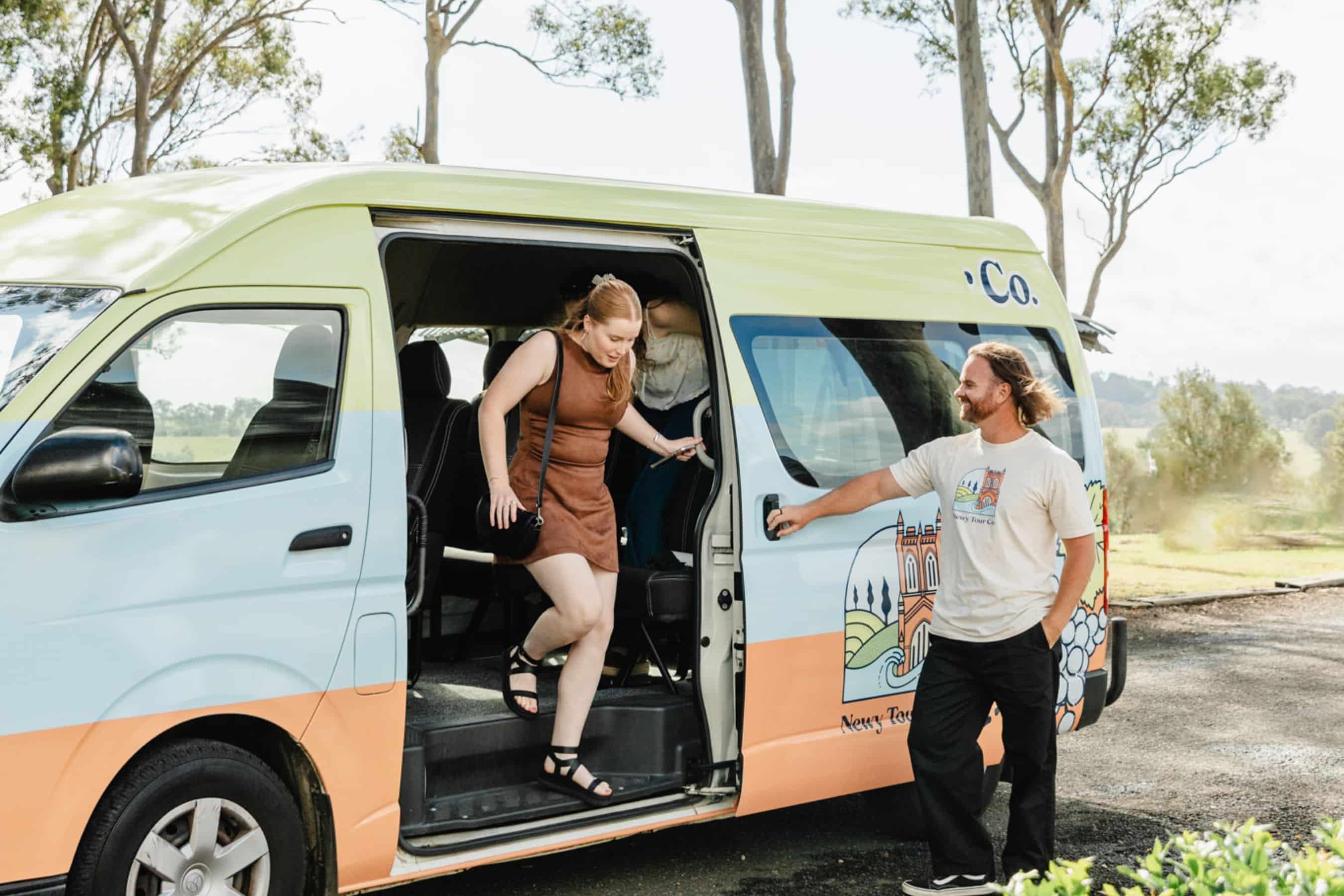 Comfortable Newy Tour Co. van with scenic outdoor backdrop, featuring friendly guide helping travelers, offering sightseeing tours in Newcastle, Australia.