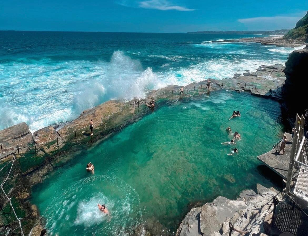 Scenic coastal pool along the Newcastle shoreline with visitors enjoying the water and ocean waves crashing nearby, offering a unique seaside experience.