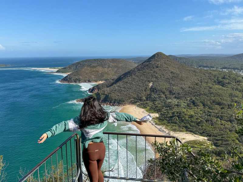 Breathtaking coastal view from a scenic lookout featuring a woman enjoying the panoramic landscape of Newy's beaches and lush green hills, perfect for tourists and adventure seekers.