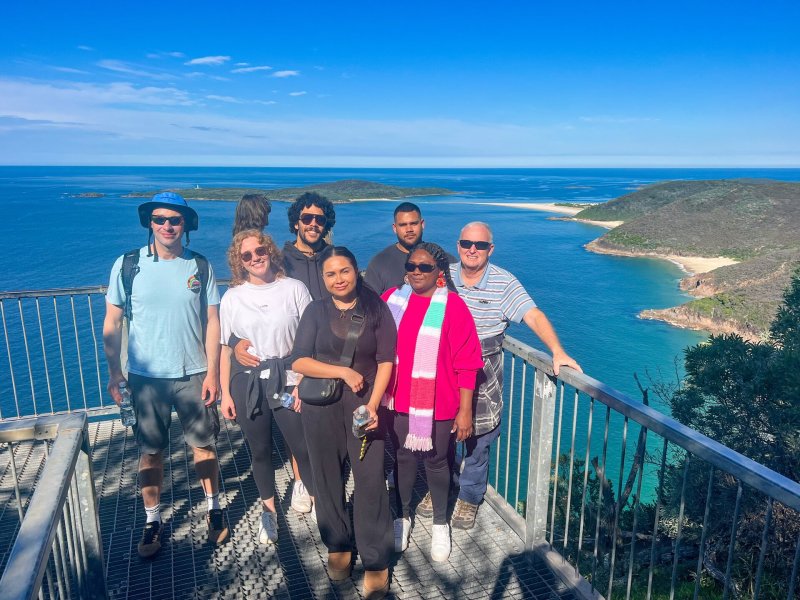 Group of tourists enjoying panoramic views of Port Stephens from a lookout, with clear blue skies and ocean in the background, during a private day tour with Newy Tour Co.