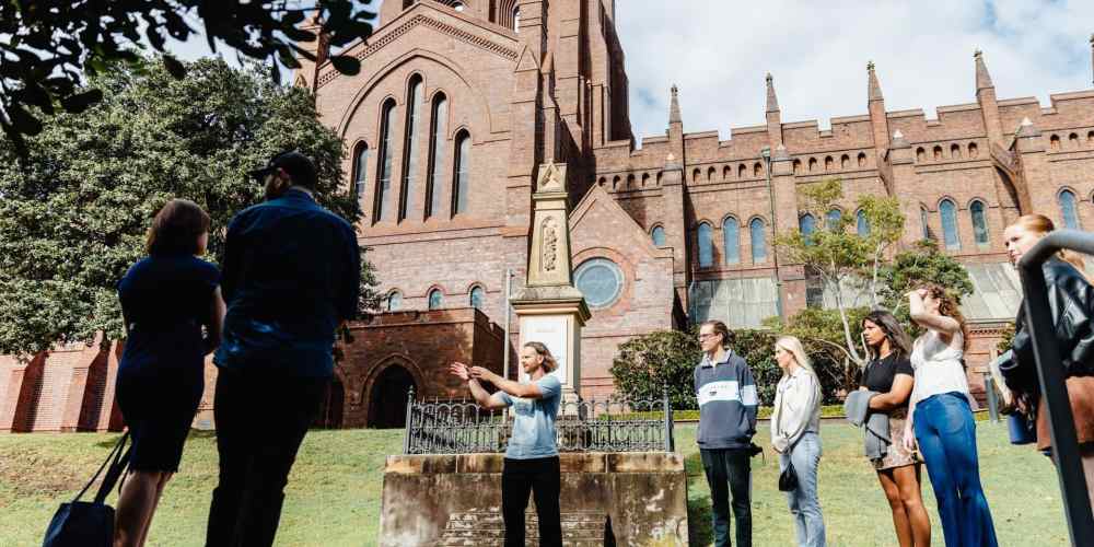 Historical city tour at Newcastle's iconic Christian missions, led by a knowledgeable guide, with visitors listening attentively in front of a stunning brick church.