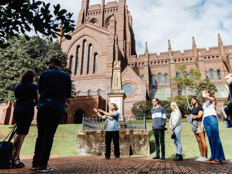 Historical city tour at Newcastle's iconic Christian missions, led by a knowledgeable guide, with visitors listening attentively in front of a stunning brick church.
