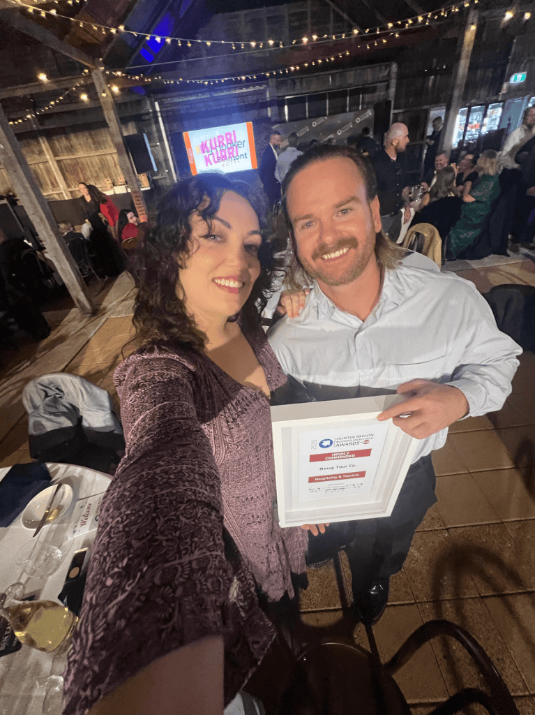Friendly couple celebrating at a Newy Tour Co. event, holding award certificate in a rustic venue with string lights, showcasing hospitality and tourism excellence in Newcastle, Australia.