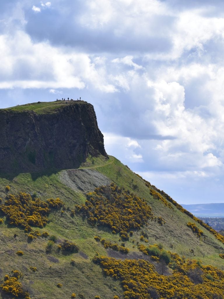 A grassy hill with yellow flowers and people standing on the cliff under a partly cloudy sky, showcasing one of the best attractions Edinburgh has to offer.