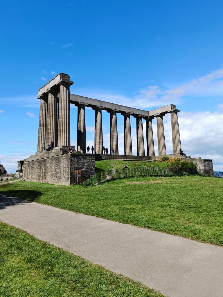 People stand by the columns of the National Monument of Scotland on a sunny day with blue sky, enjoying one of the best attractions Edinburgh has to offer.