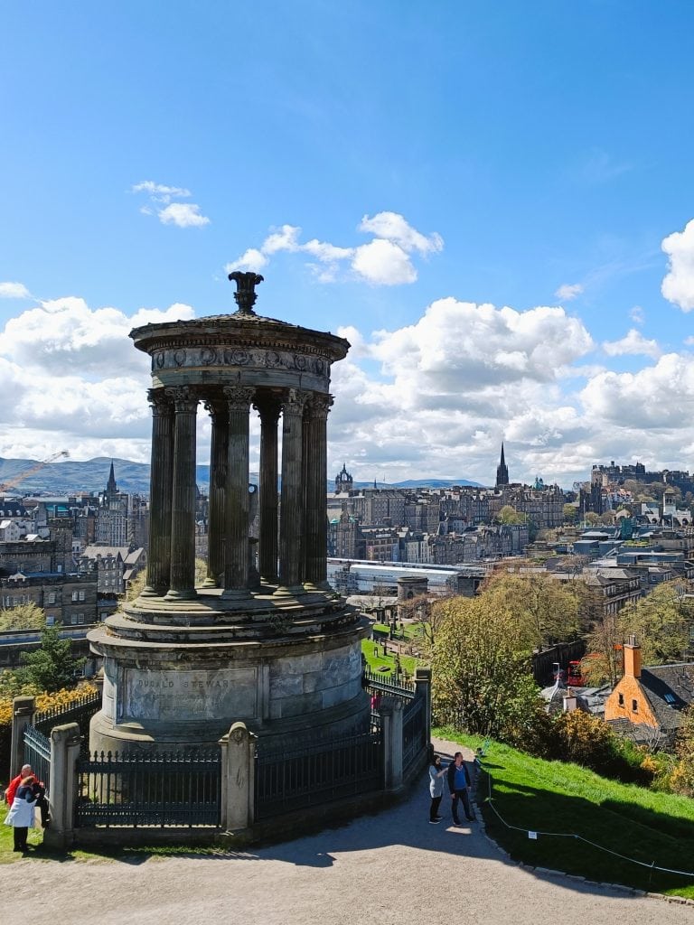 View of the Dugald Stewart Monument overlooking Edinburgh city, one of the best attractions Edinburgh has to offer, under a bright blue sky with scattered clouds.