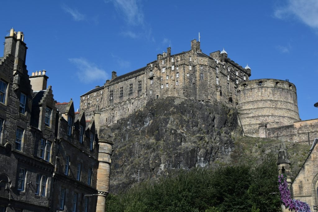 Edinburgh Castle, one of the best attractions Edinburgh has to offer, sits atop a rocky hill under a blue sky, with historic buildings in the foreground.