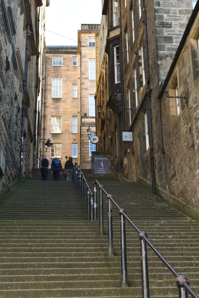 Wide stone staircase between tall buildings, with a few people walking up and handrails on both sides—one of the best attractions Edinburgh has to offer for those exploring the city's charming old streets.