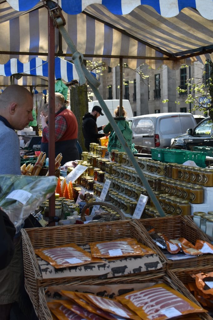 Outdoor market stall with jars of honey, packaged meats, and people browsing under a striped canopy—one of the best attractions Edinburgh has to offer.