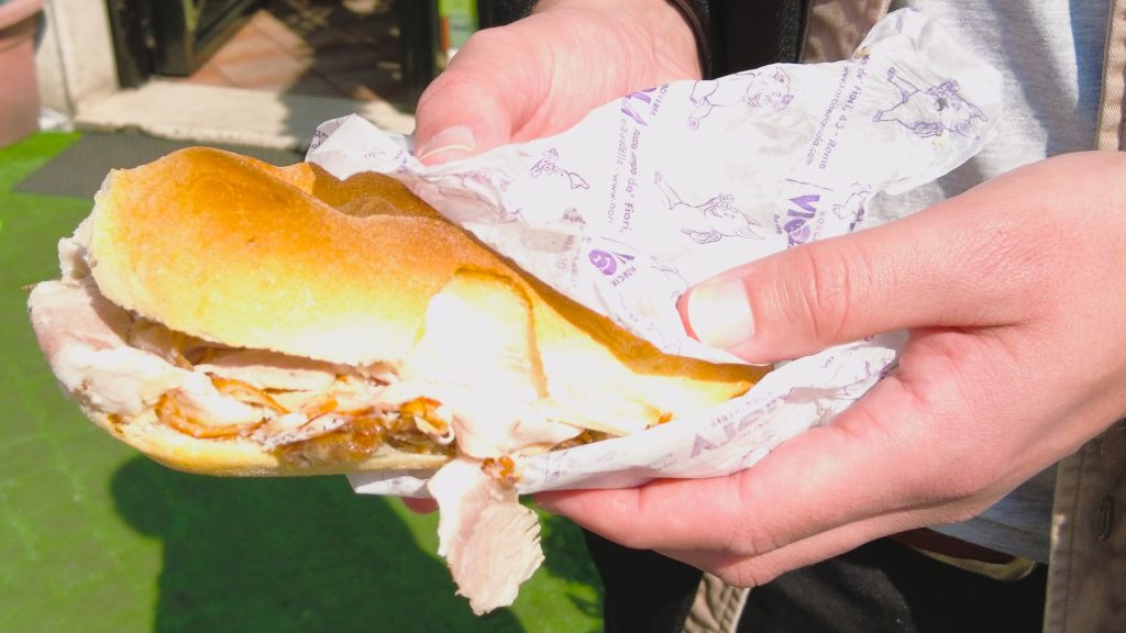 Person holding a must-try sandwich with sliced meat in a paper wrap on a sunny day, offering a delightful taste of Italian food.