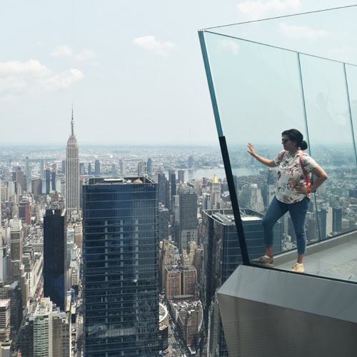 Person standing on a glass ledge overlooking a cityscape with skyscrapers and a cloudy sky.