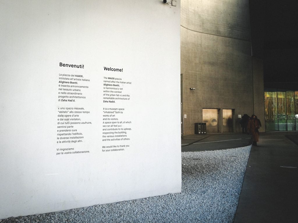 A modern entrance at the MAXXI museum in Rome features welcome text in Italian and English on a wall, with visitors exploring in the background.