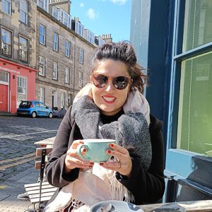 Woman sitting outdoors at a cafe, smiling, holding a mug, wearing sunglasses and a scarf.