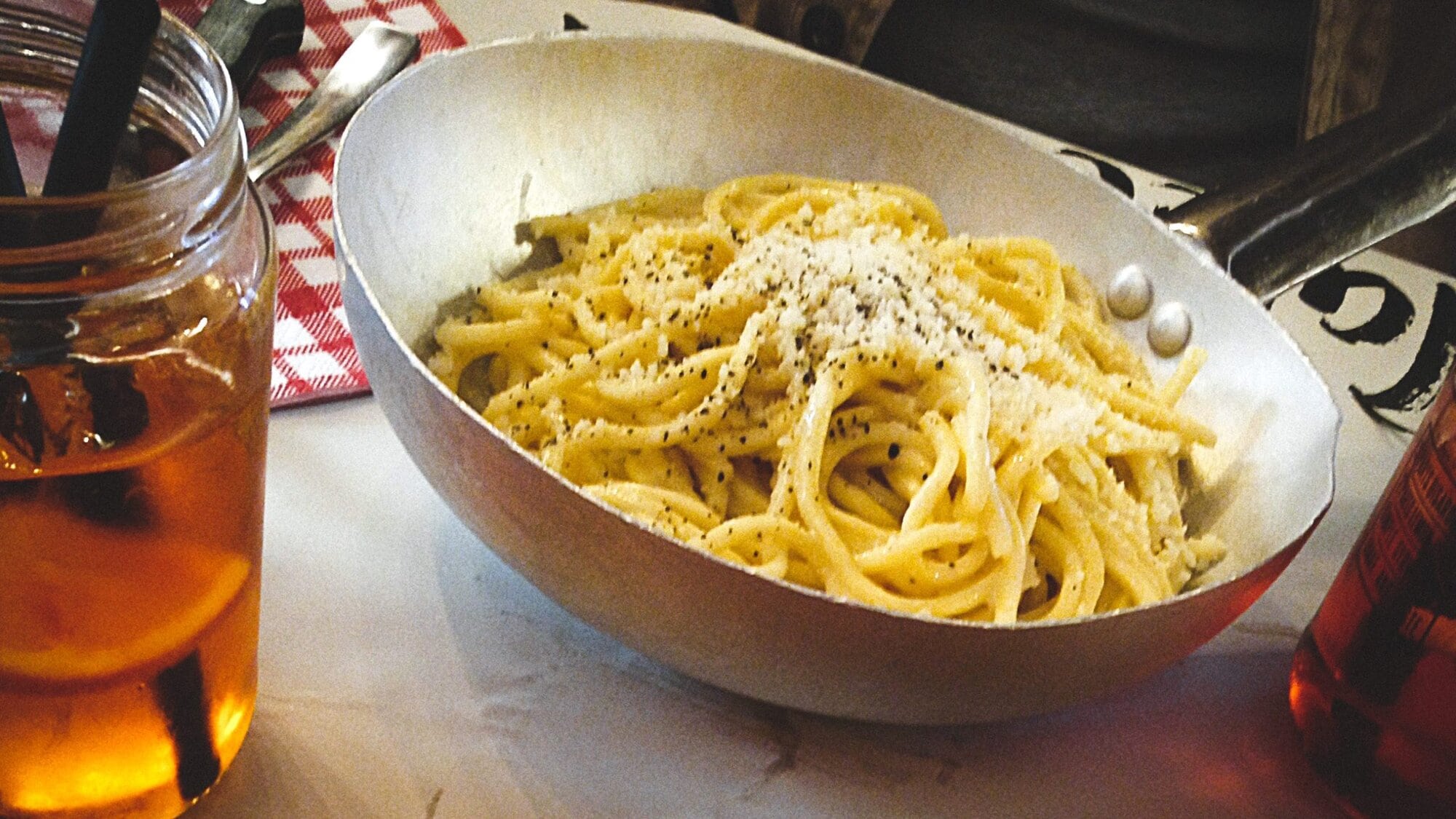 This must-try Italian food features pasta with cheese and pepper in a metal pan, surrounded by enticing drinks and a plate of cured meat.