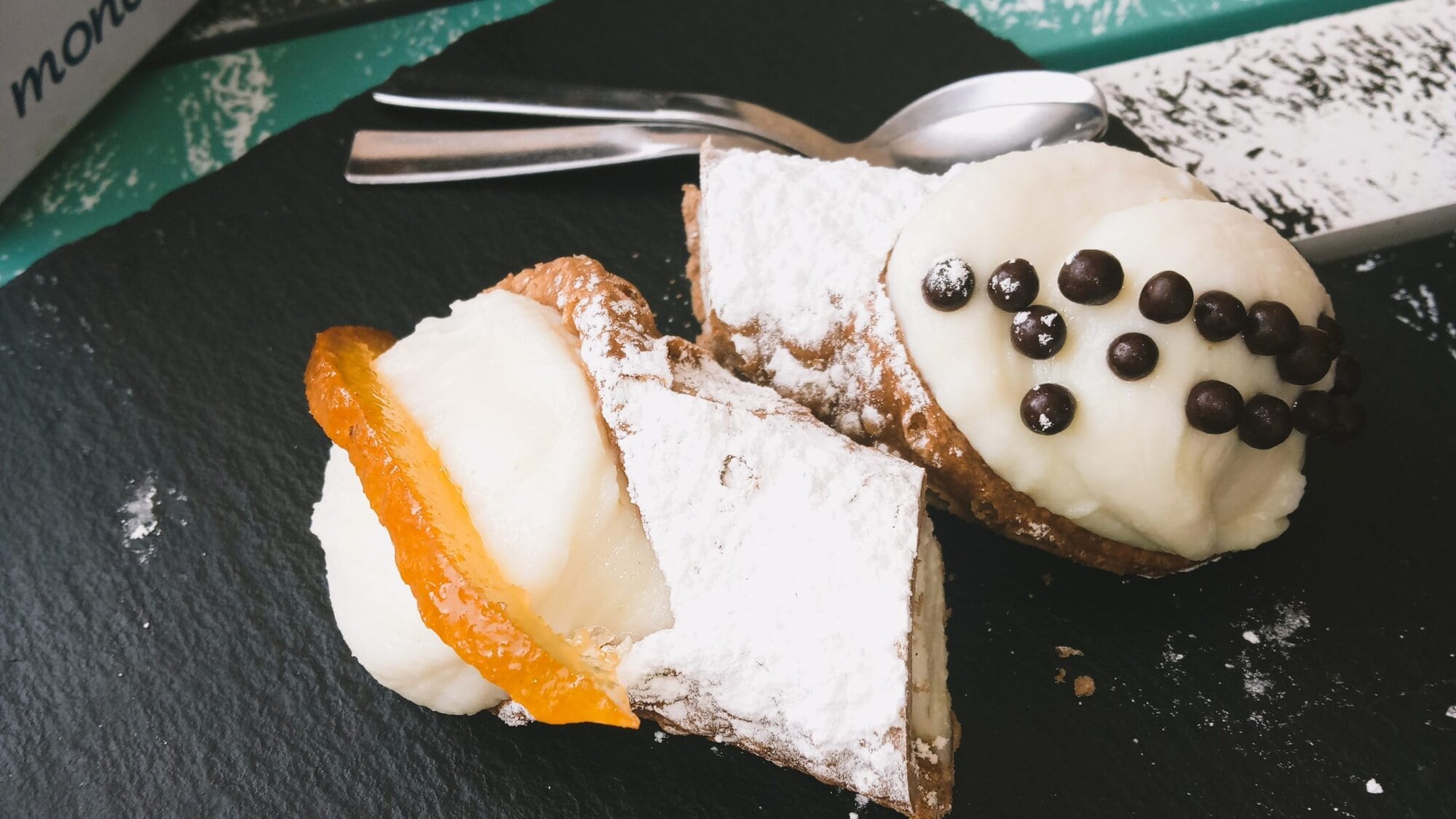 A must-try Italian food delight: cannoli and a chocolate-dotted pastry rest elegantly on a slate platter atop a colorful table with a branded tabletop display.