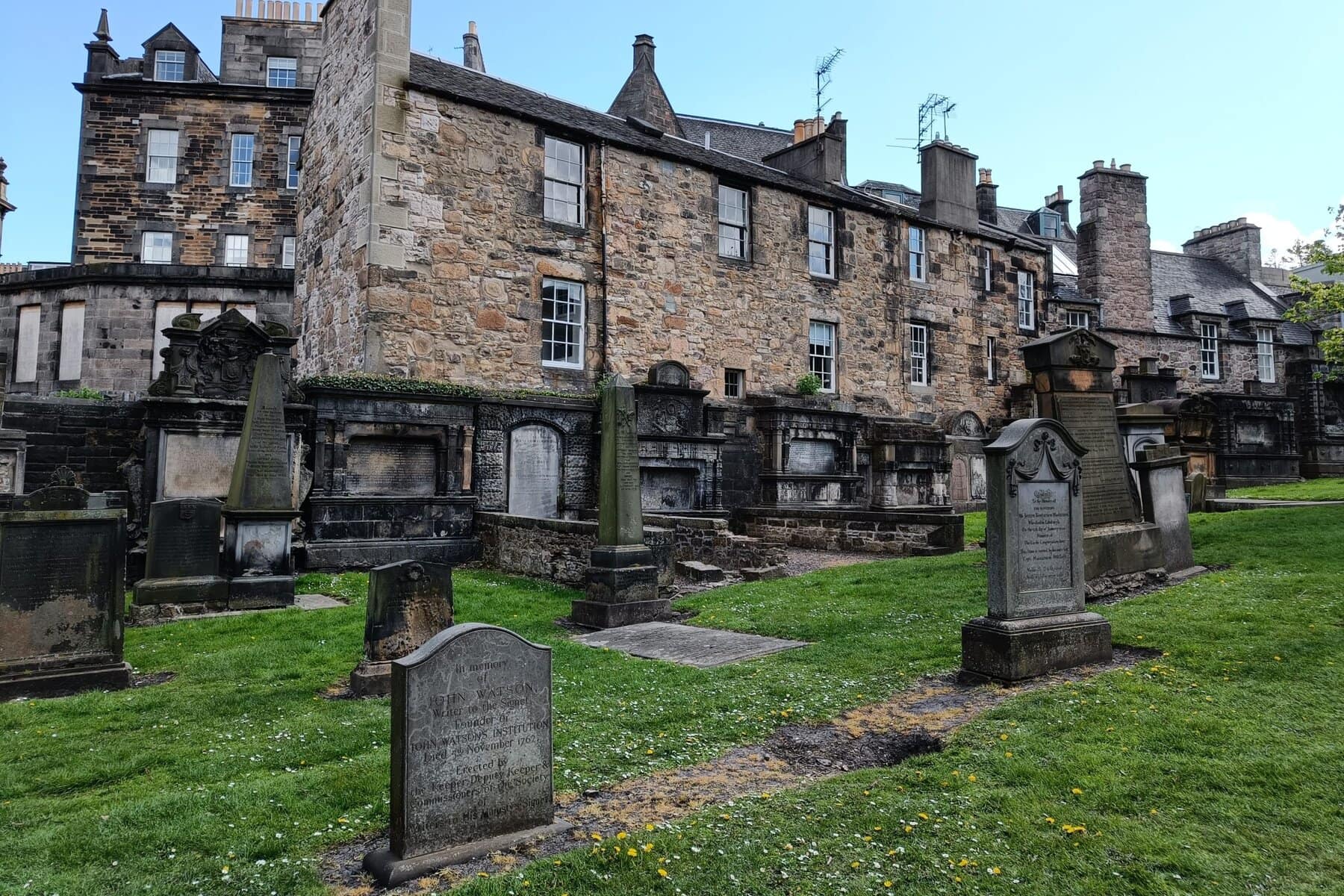 Old gravestones and stone buildings in a historic cemetery with grass and blue sky, showcasing one of the best attractions Edinburgh has to offer.