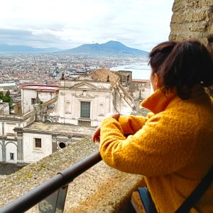 A woman in a yellow coat looks out over Naples with Mount Vesuvius in the distance.