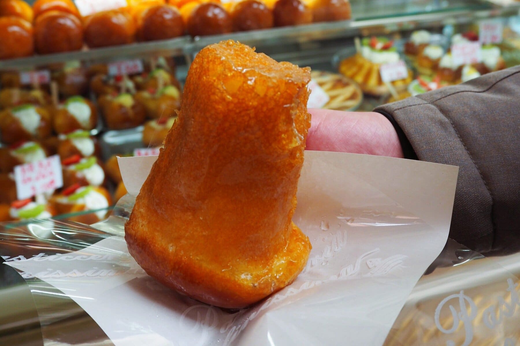 A hand holds a glazed pastry wrapped in paper in front of a bakery display filled with assorted desserts—a must try food showcasing the delights of Naples cuisine.