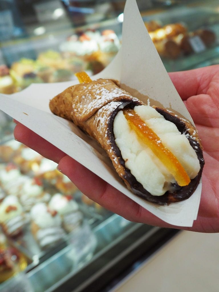 A hand holding a Sicilian cannoli with ricotta filling and candied orange on top, in a pastry shop—this treat is a must try food in Naples for anyone exploring Naples cuisine.