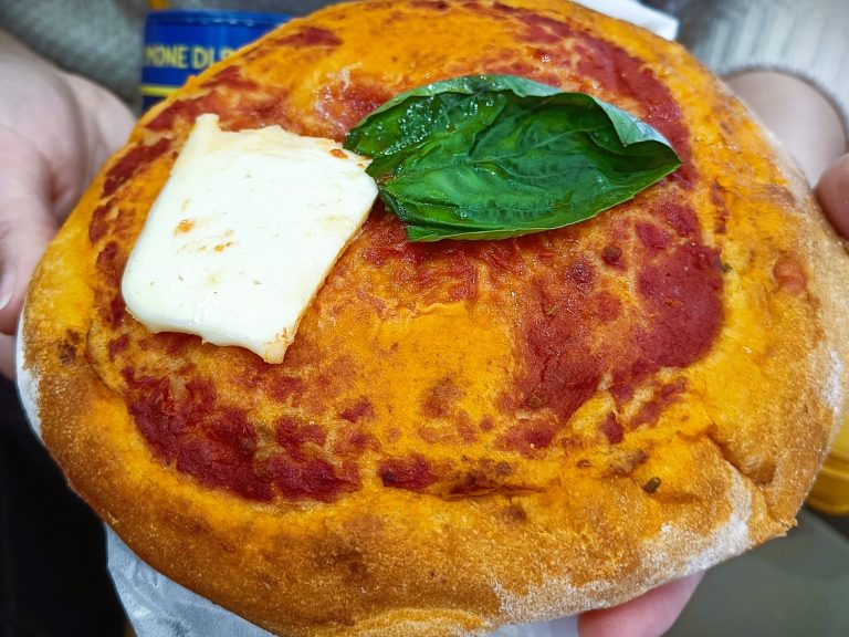 A hand holds a small pizza topped with tomato sauce, basil leaf, and a slice of melted cheese—a delicious sample of must-try food Naples is famous for.
