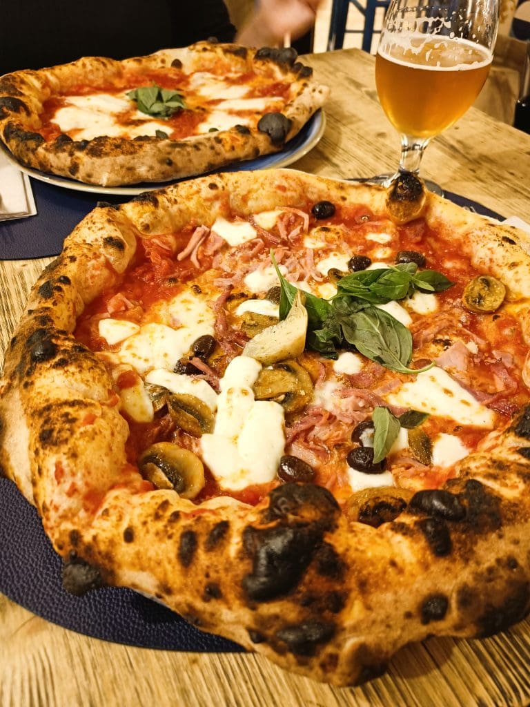 Two wood-fired pizzas with cheese, mushrooms, and basil, served with a glass of beer on a wooden table—a perfect introduction to classic Naples cuisine and must-try foods for any pizza lover.