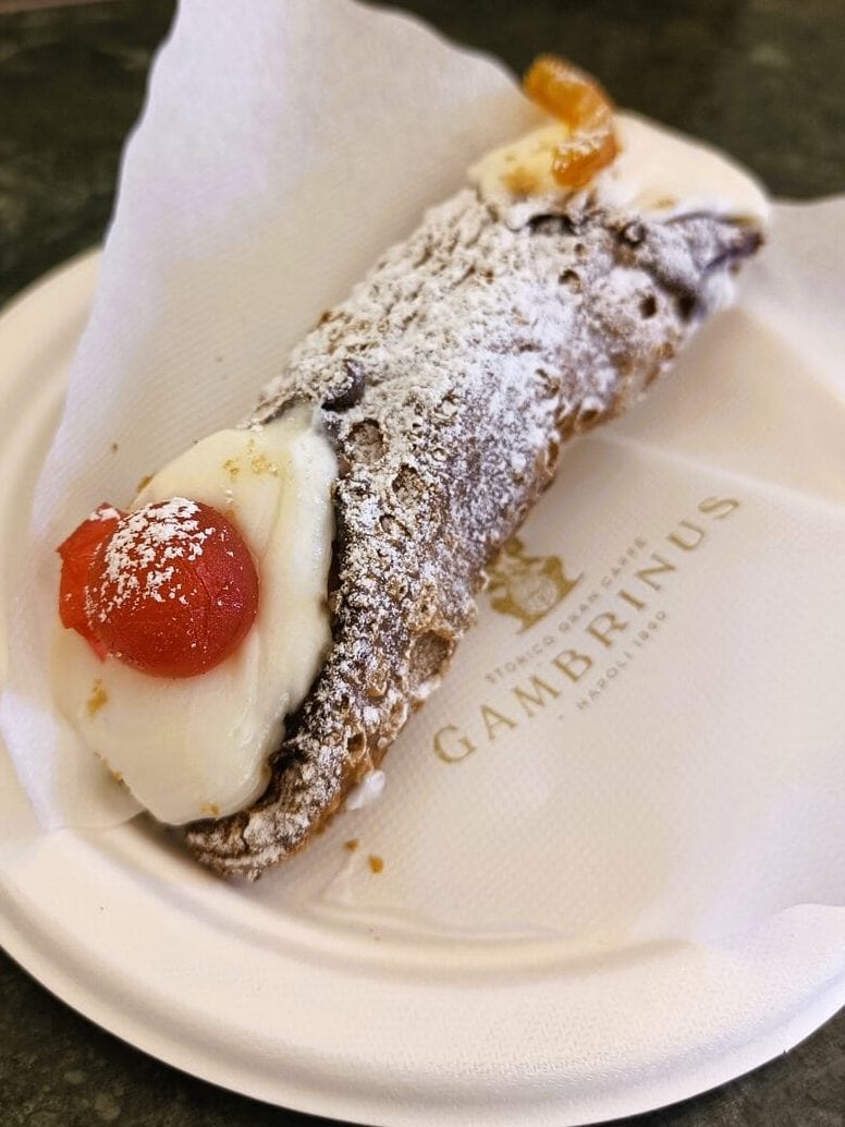 A Sicilian cannolo with ricotta filling, powdered sugar, and candied fruit on a paper plate—a sweet treat often enjoyed alongside other classic Naples cuisine.