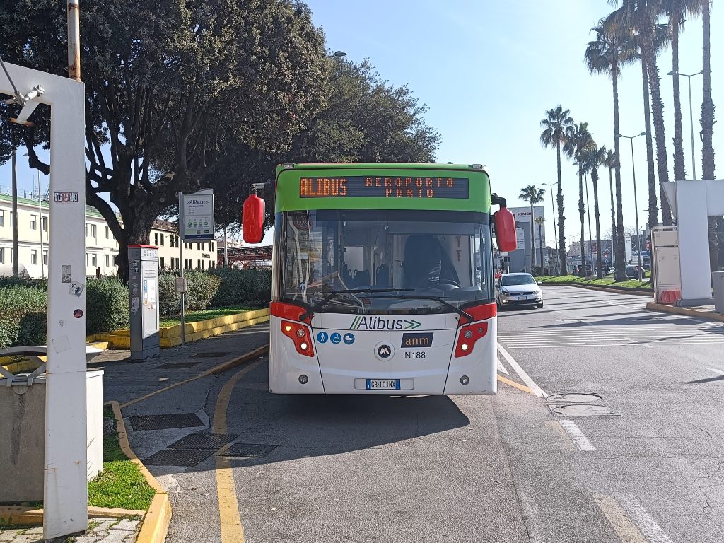 A green and white airport shuttle bus labeled "Alibus," part of Naples transportation, is parked at a sunny street stop.