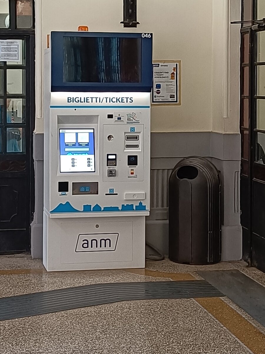 Ticket vending machine in a hallway, helping travelers get around Naples, with people and shops in the background.