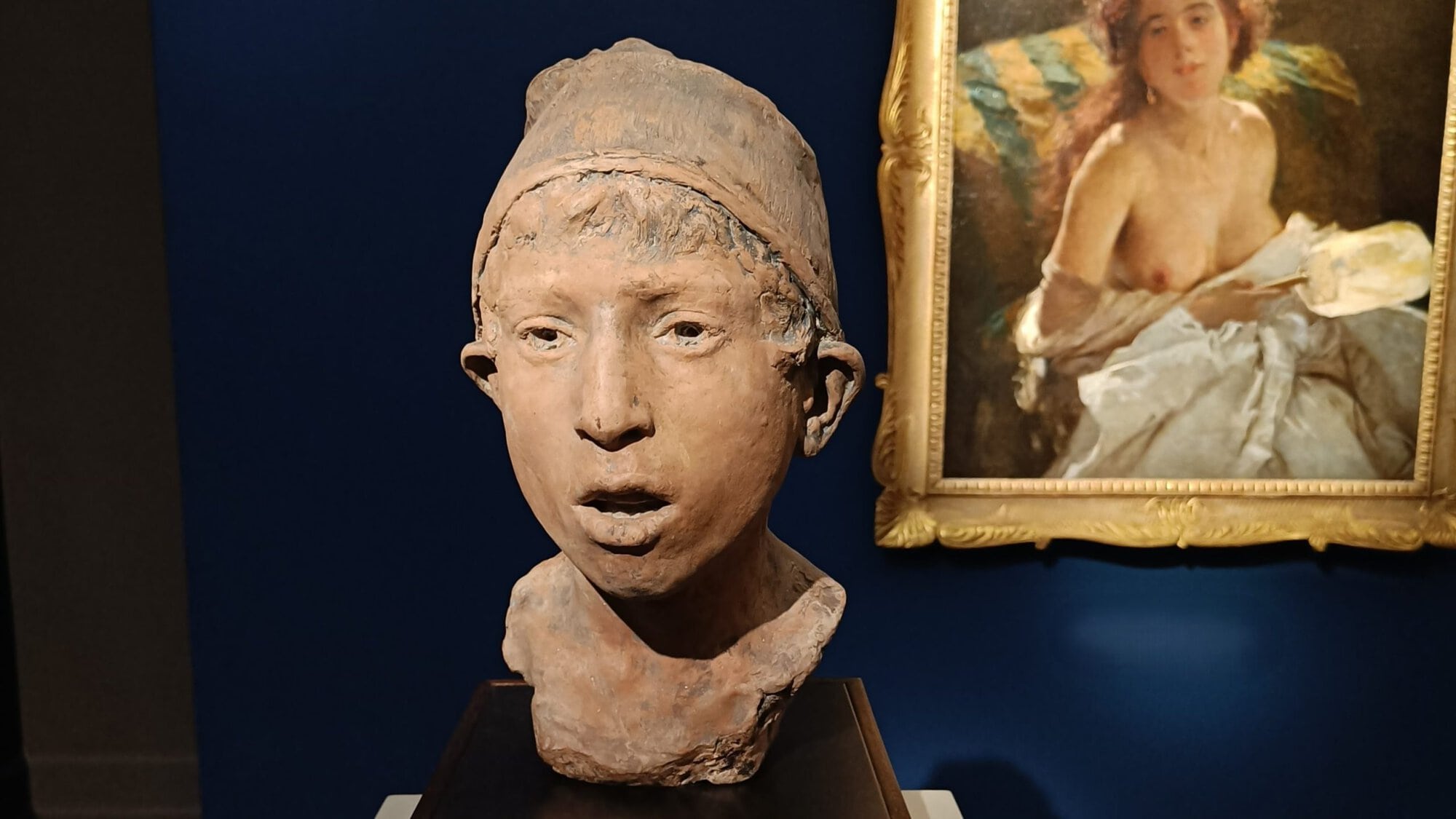 Clay bust of a young person with a cap, displayed on a pedestal; painting of a woman in the background—a scene reminiscent of classic exhibits in the best museums Naples has to offer.