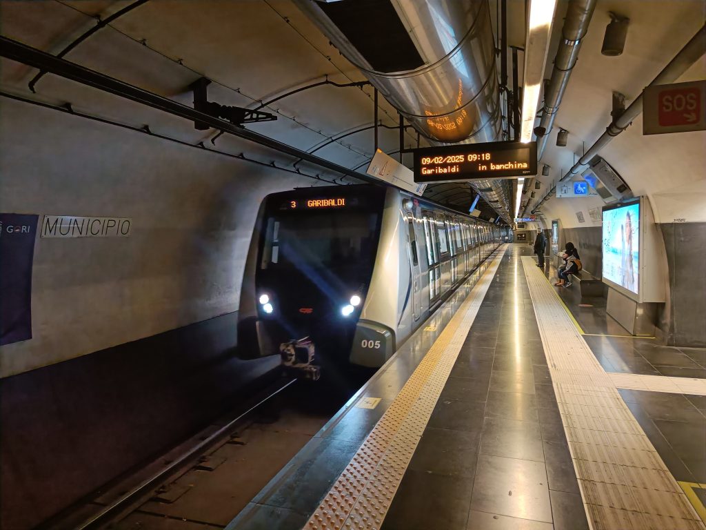 A modern subway train arrives at an underground station, offering a glimpse into efficient Naples transportation as a few people wait on the platform.