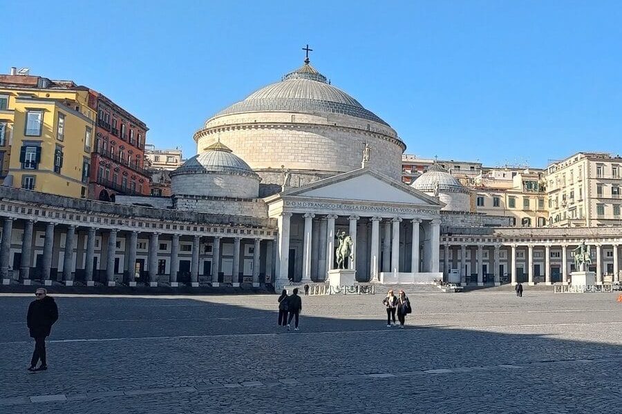 Wide plaza with people walking, a domed church, columns, and colorful buildings under a clear blue sky—this vibrant scene captures the essence of one of the popular neighborhoods in Naples.