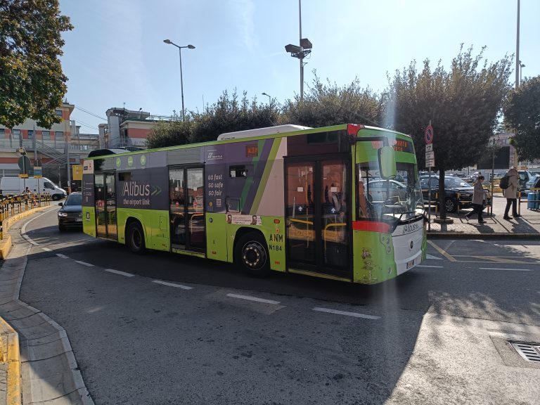 A green city bus labeled "Alibus" drives on a sunny street near a sidewalk with trees and people, offering a convenient way for travelers searching for where to stay in Naples or looking for nearby accommodation in Naples.