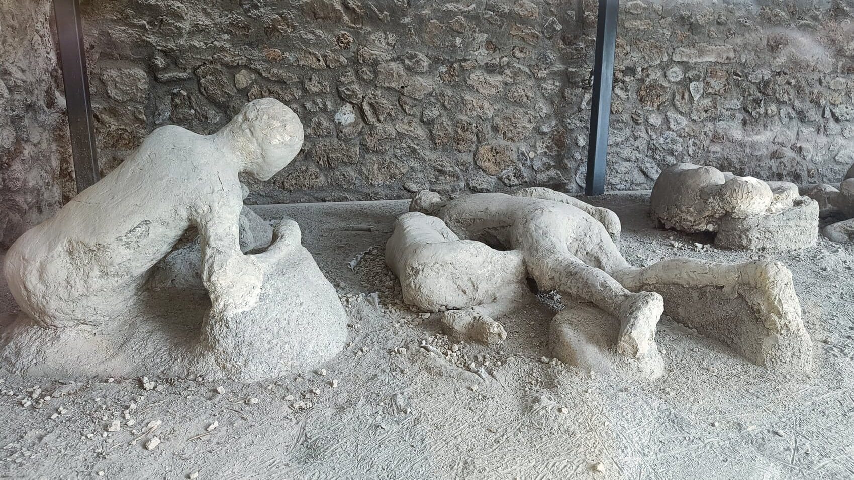 Plaster casts of human figures preserved in ash at an archaeological site with a stone wall in the background, as seen in some of the best museums in Naples.