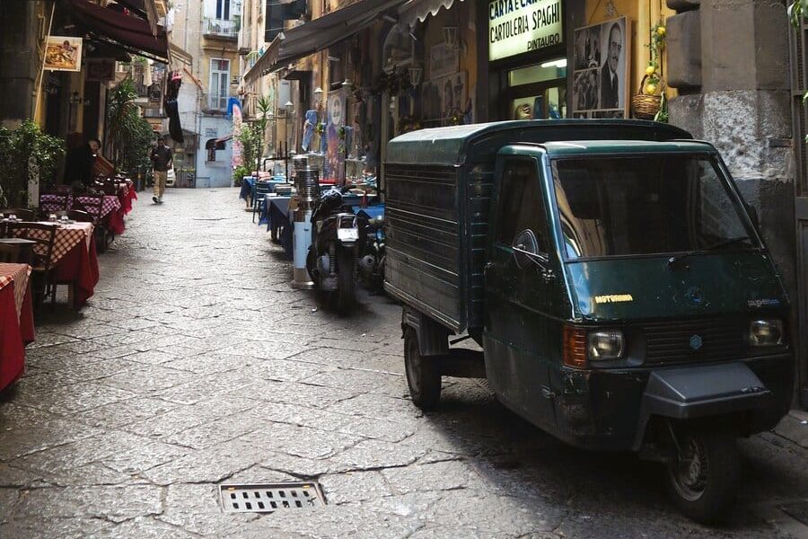 Narrow cobblestone street in one of the popular neighborhoods of Naples, with parked scooters and small vehicles beside outdoor cafes and old buildings, showcasing the lively charm typical of Naples neighborhoods.