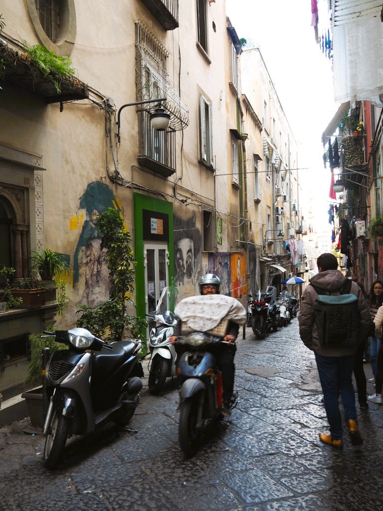 Narrow cobblestone street with scooters, pedestrians, old buildings, and murals—a classic scene among the many Naples attractions and a must-see for those seeking authentic things to do in Naples.