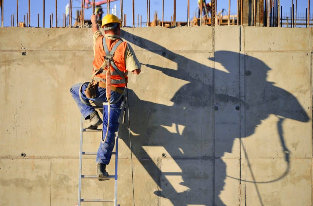 A construction worker climbs a ladder, casting a shadow on the concrete wall at a building site.