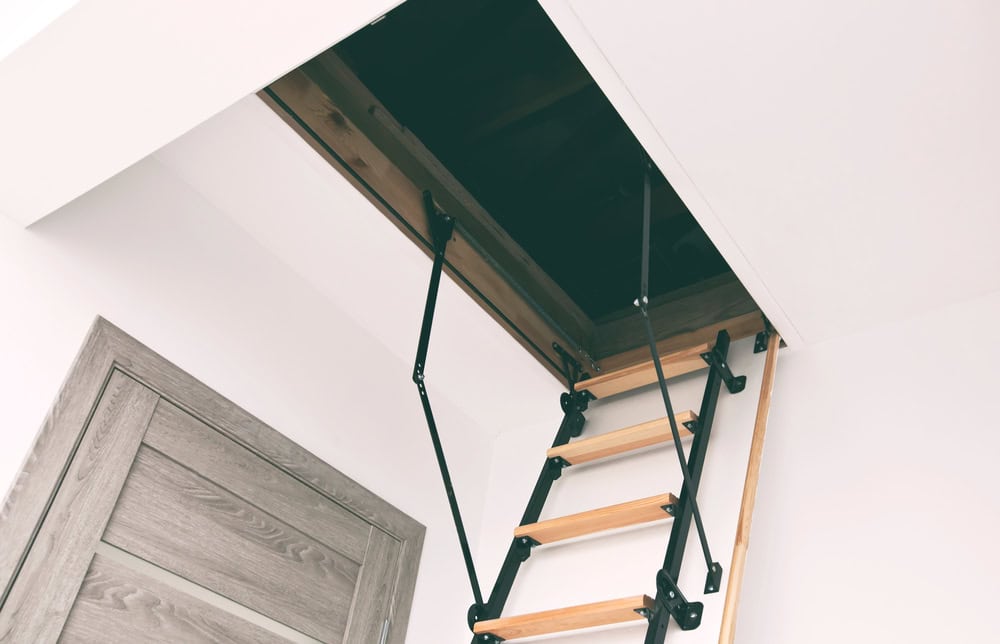 Folding attic ladder extending from a ceiling hatch, combining sleek black metal hardware with natural wooden steps