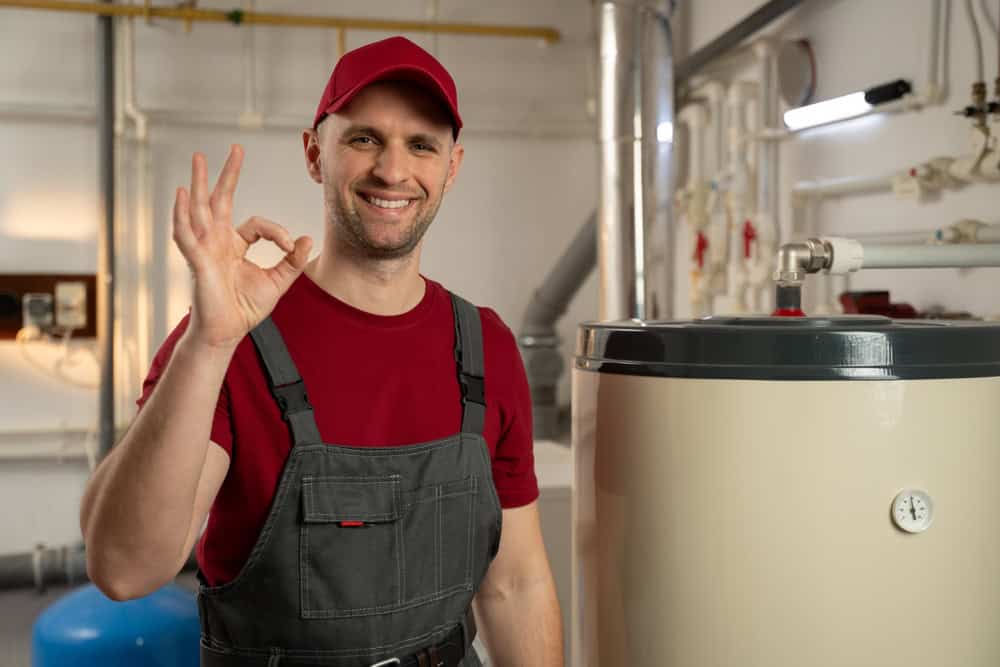 Maintenance professional in red cap and overalls gives OK sign while standing next to water heater system in utility room