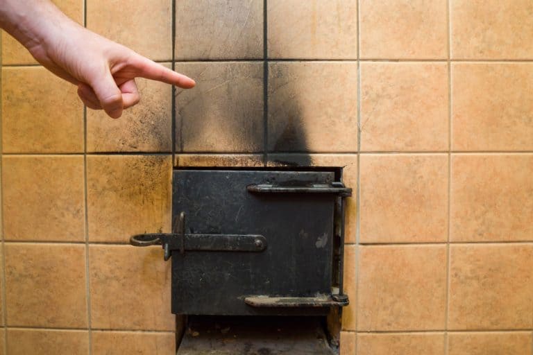 A hand points to a small, blackened oven door set into a tiled wall.