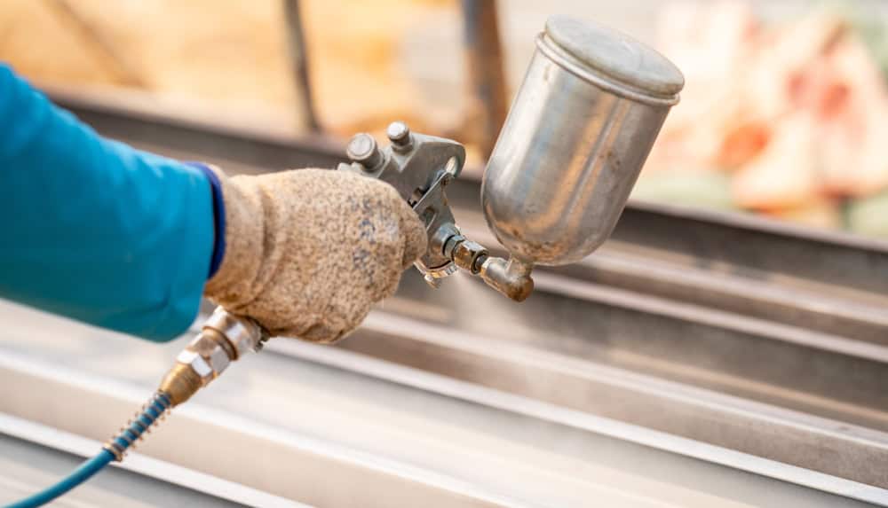 A worker in gloves uses a spray gun to evenly coat metal sheets with paint.