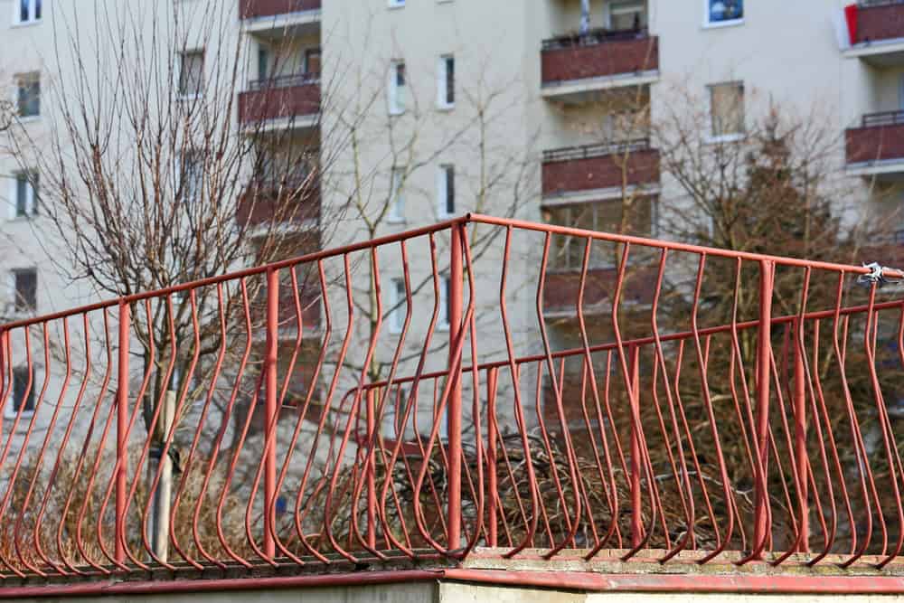 A red, bent iron fence stands in front of a residential apartment building, with leafless trees signaling winter's arrival.