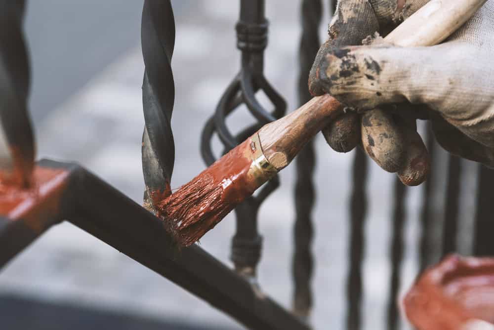 A gloved hand carefully paints a wrought iron fence with a fresh coat of rust-red paint.