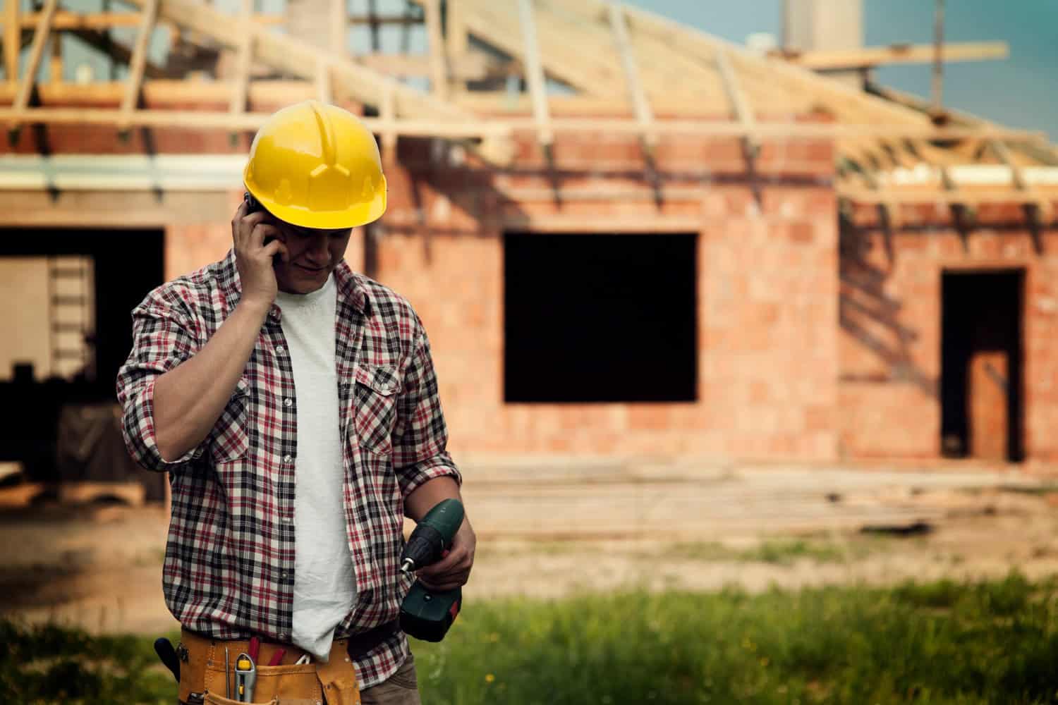 Construction worker in plaid shirt and yellow hard hat takes a phone call while holding a power drill at a residential building site