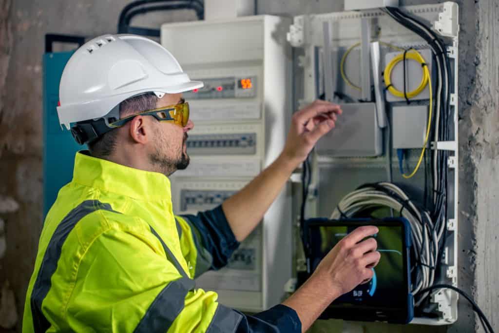 Electrical inspector in safety gear examines a circuit breaker panel while holding a tablet device, demonstrating modern safety protocols and technological integration in electrical maintenance.