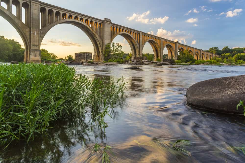A serene river flows beneath an elegant stone viaduct, framed by lush greenery and a clear blue sky.