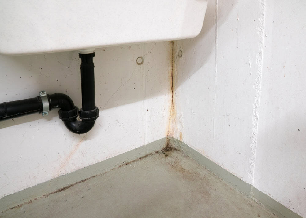 Rusty stains and grime accumulate around the sink's plumbing in a neglected bathroom corner.