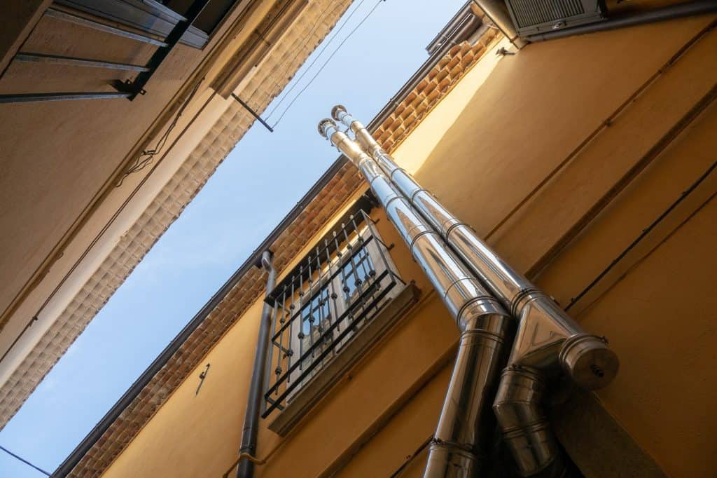 Metal exhaust pipes rise along a yellow-painted exterior wall, beneath a wrought iron balcony railing and terracotta roof tiles, creating an industrial contrast against the traditional architecture.